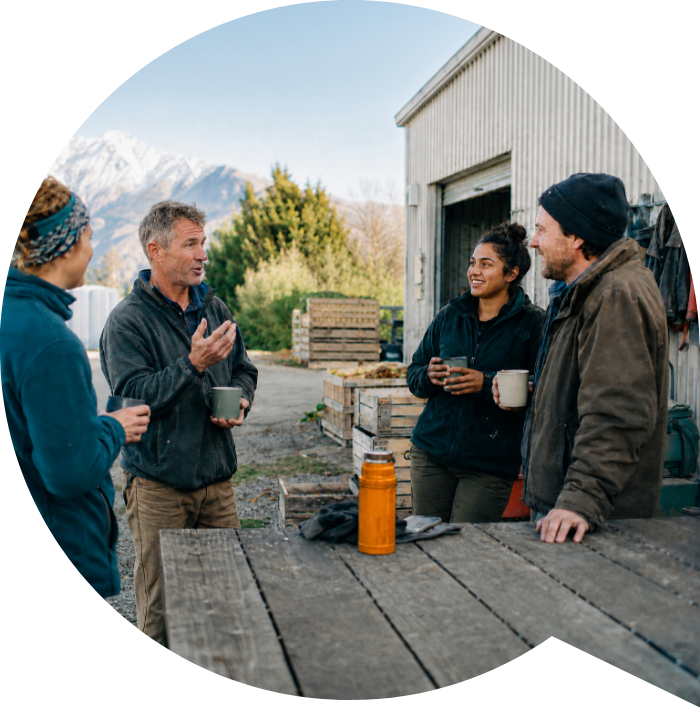 A small team gathered together outside a rural workshop, talking over coffee.